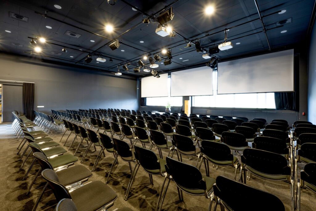 Moderne conferentieruimte aan de Johan Huizingalaan met rijen zwarte stoelen en grote projectieschermen.